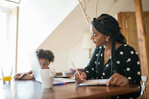 Single mother on Laptop at kitchen table while child does homework beside her.