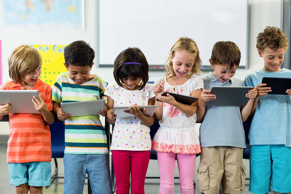 A row of kids stand in front of a classroom holding tablets.