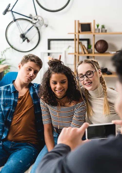 A boy takes a picture of his teen friends on a living room couch.