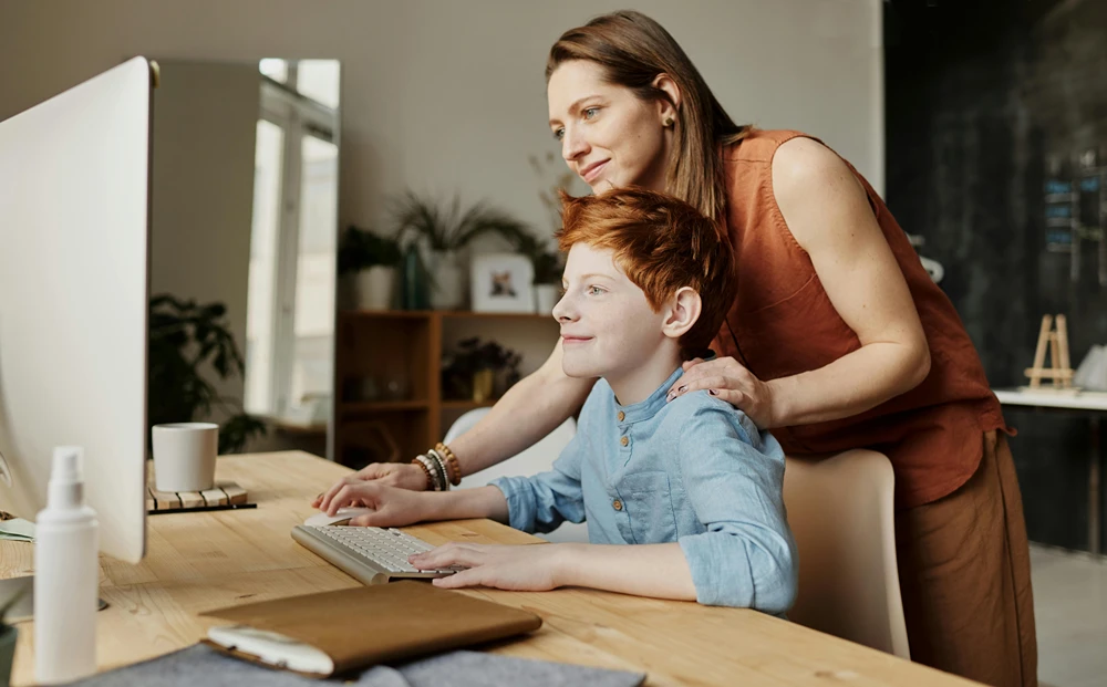 Woman looking over boy’s should as he uses a computer.