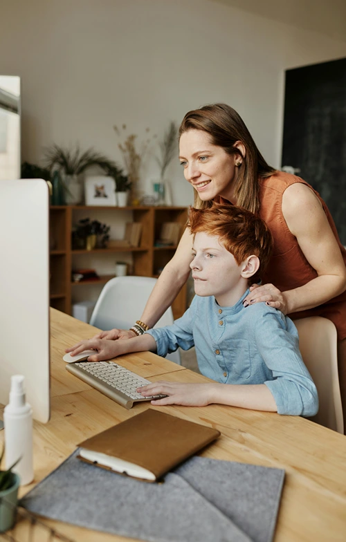 Boy using computer while his mother helps guide the mouse.
