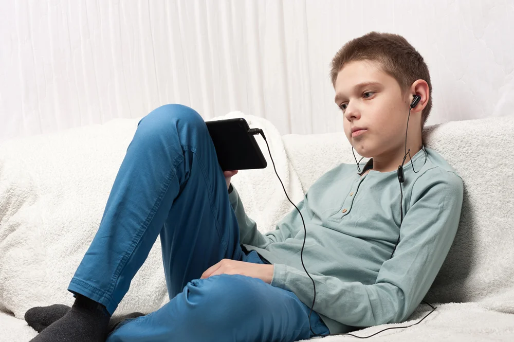A teen boy girl is laying back and listening to an audiobook on tablet.