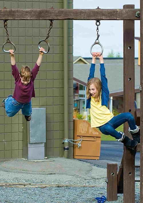 A girl and boy hanging on rings in a school playground.