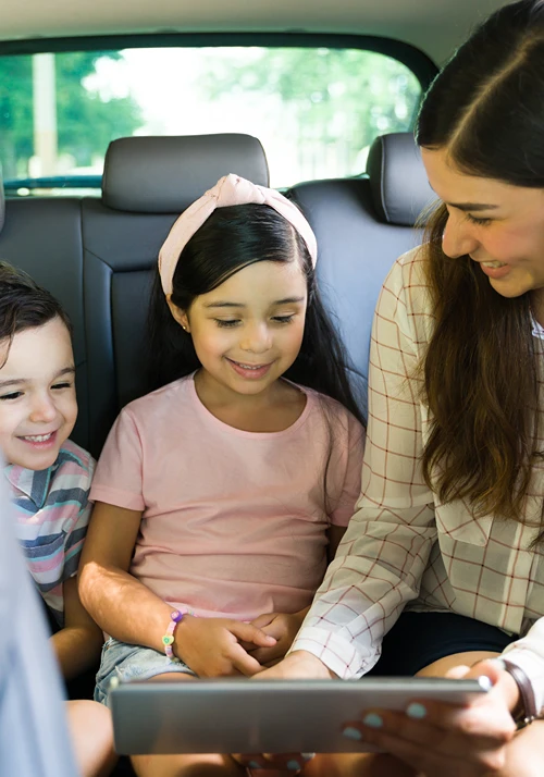 Mom showing tablet to her children in back seat of car before a road trip.