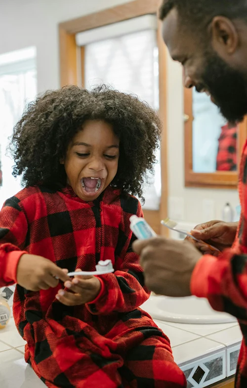 A dad puts toothpaste on a brush held by his daughter.