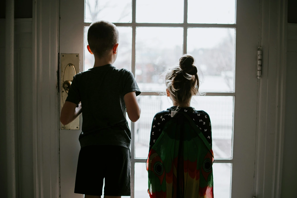 A boy and a girl standing and looking out the window of their front door.