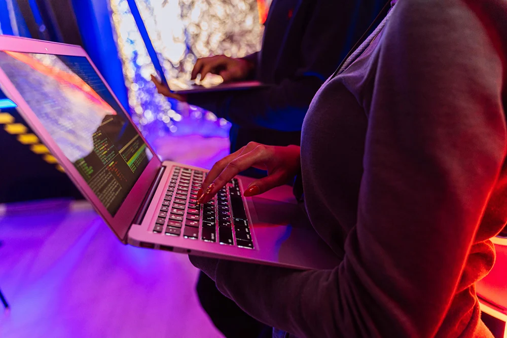 Two people standing while holding and typing on their laptops.