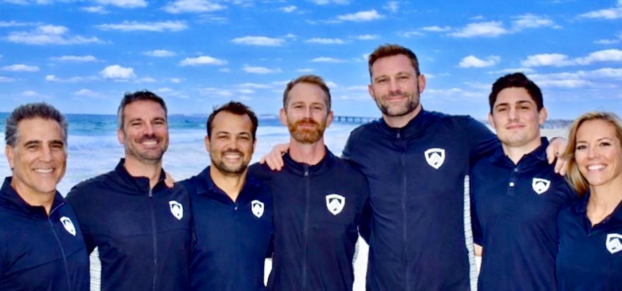 Seven team members of Higher Grounds Mgmt smiling in matching navy uniforms with the company shield logo, standing arm in arm on a sunny beach.