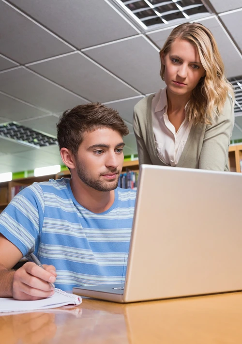 Tutor stands above seated male with pen and paper in right hand and laptop on his left.