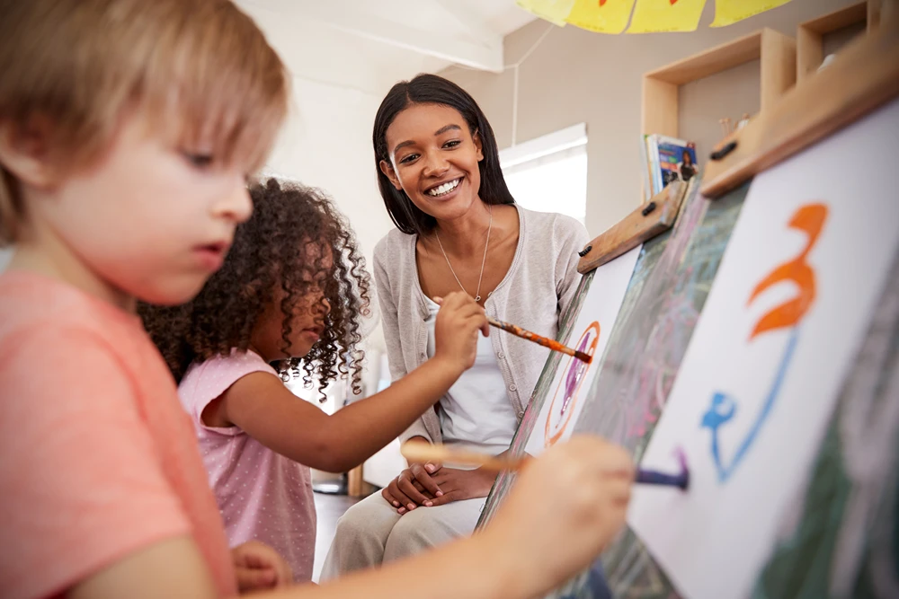 A smiling art teacher watches her young students painting on easels.