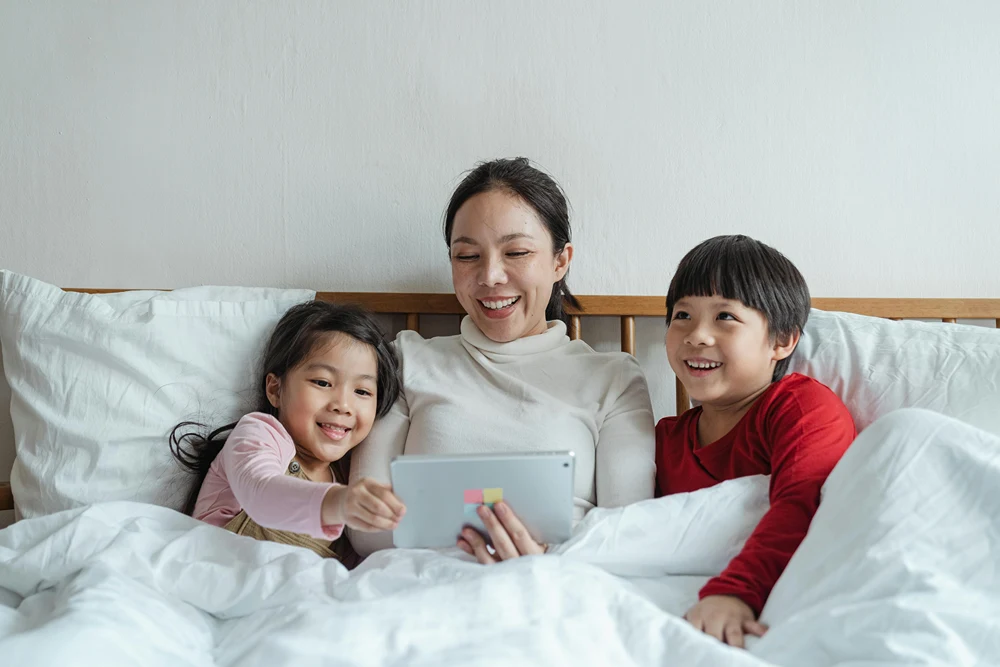Mom in sitting up in bed with tablet, as her son and daughter on both sides look at the screen and smile.