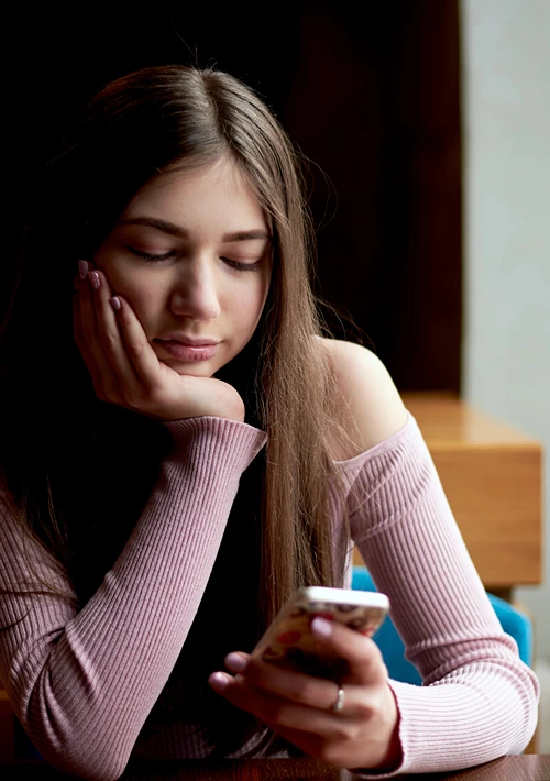 A teenage girl rests her chin in one hand, eyes fixed on her smartphone held in the other, completely absorbed in what she’s seeing.
