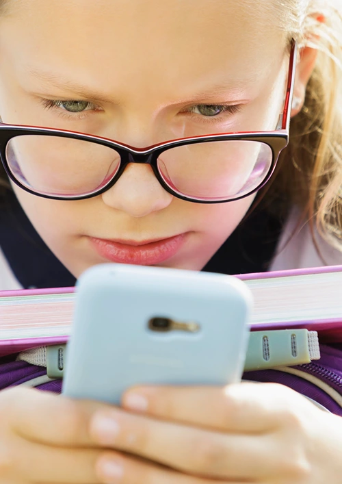 A girl with glasses holding a text book while looking closely at her smartphone.