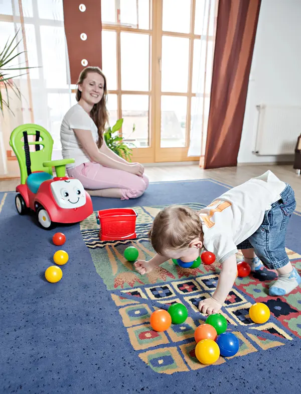 Toddler plays on the floor with toys as mother watches.