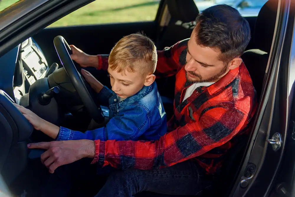 Boy sits on father's lap in front seat of can learning the features on the dashboard.