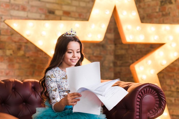 Girl dressed wearing a tiara script backstage with big star behind her. 