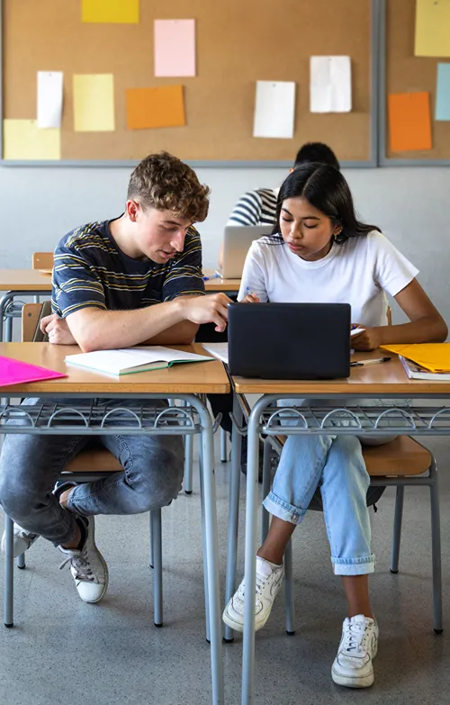 Two teens with desks pulled together in classroom.