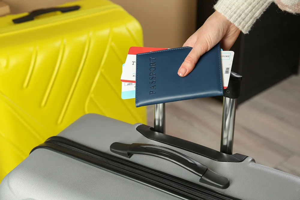 Hand of a woman rolling her luggage while holding tickets and passport.
