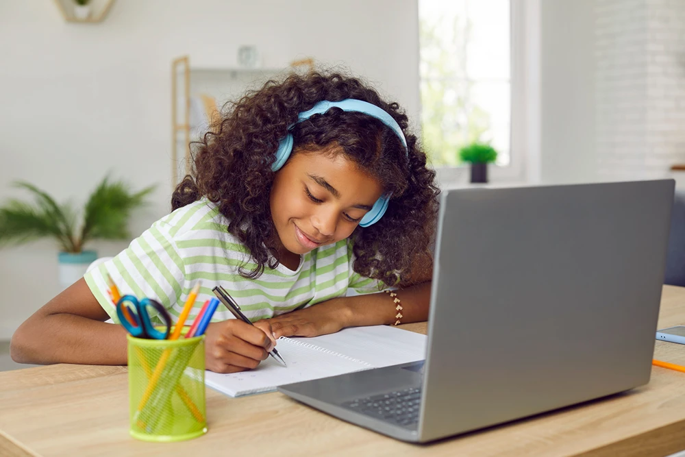 A girl wearing headphones is writing on a note pad in front of her laptop.
