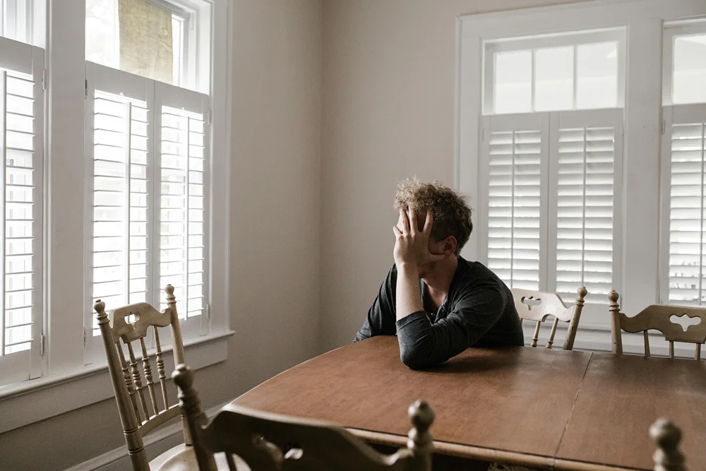 A distraught man with hand on his head sitting at kitchen table.