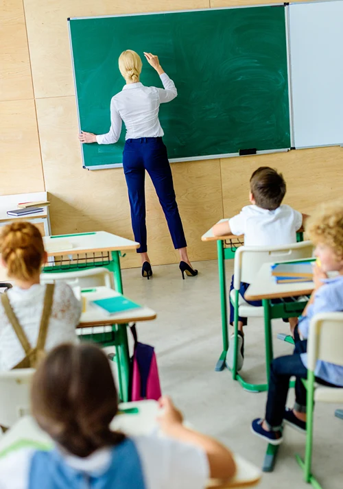 Teacher writes on greenboard in front of classroom full of kids in desks.