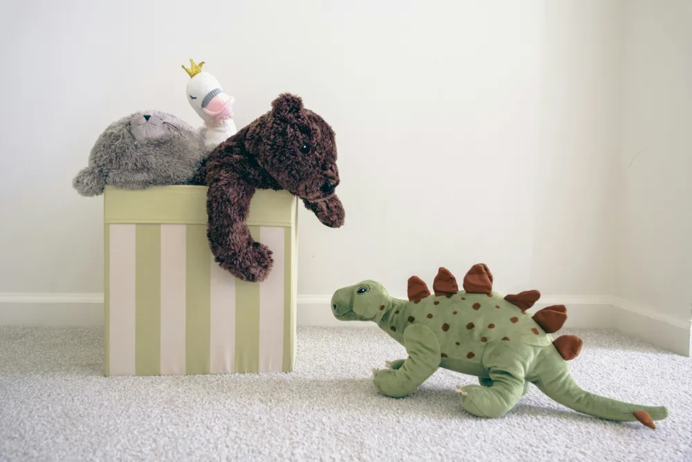 A stuffed bear looking out from inside a box at a stuffed dinosaur. 