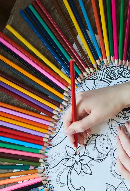 Coloring page surrounded by pencil crayon as hand works on it.