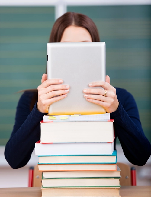 An student hides her face behind a pile of books and a tablet.
