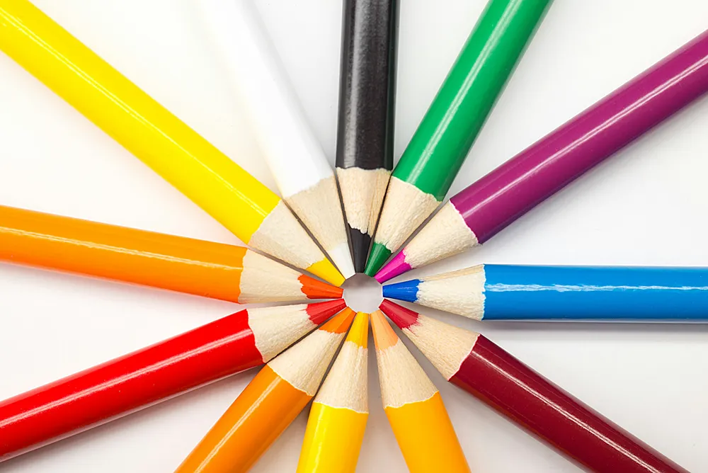 Close-up of sharpened colored pencils arranged in a rainbow circle with tips pointing inward, creating a star-like pattern on a white background. Perfect for themes of creativity, art, school supplies, and stationery.