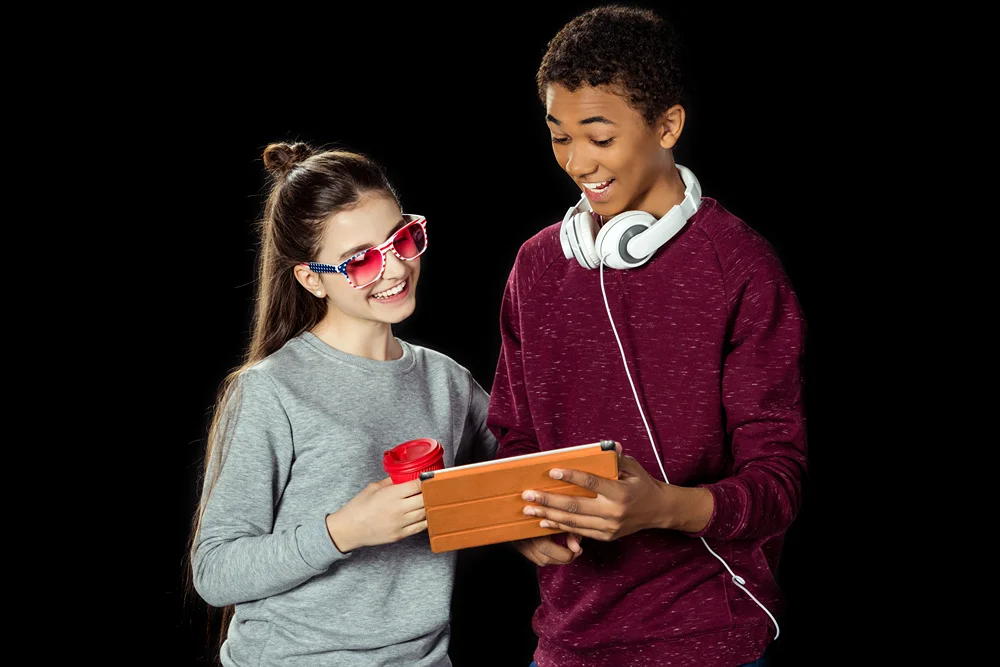 Two smiling teenagers using a tablet together, girl holding a red cup and wearing sunglasses, boy with headphones around his neck.
