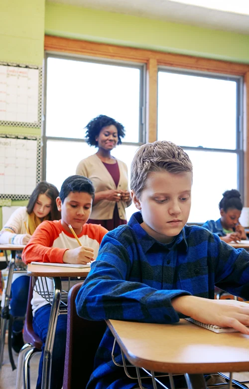 Kids in classroom at desks with teaching in the background.