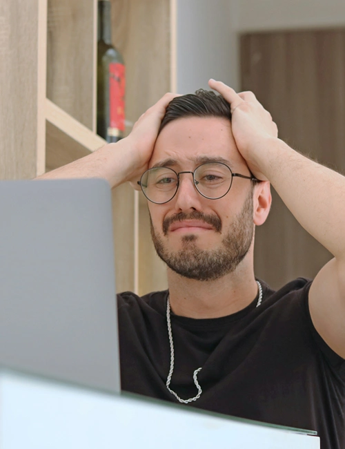 A male student holds both hands on his head as he distressingly stares at his laptop screen.