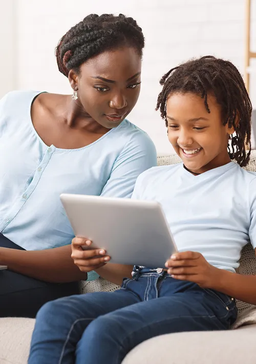 A mother site on sofa with her daughter who is using a laptop.