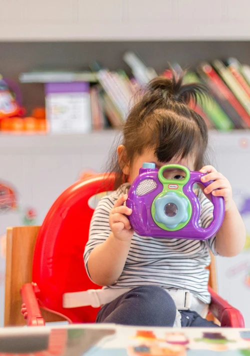 Pre-school girl sitting at a table looking through the handle of a toy at the camera.