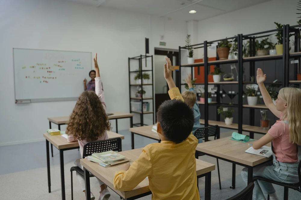 Kids holding up their hands as their teacher writes on a whiteboard.