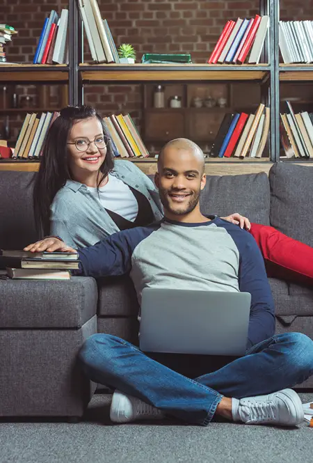A woman on a couch with books and a man seated on floor with a laptop.