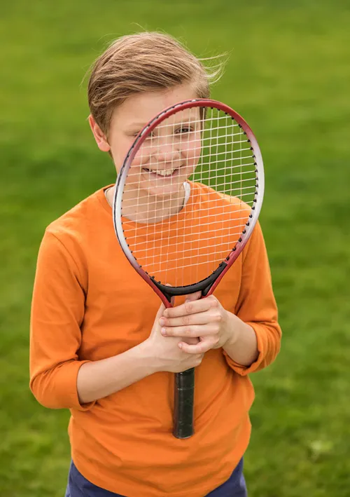 Boy outside smiling through his tennis racket.