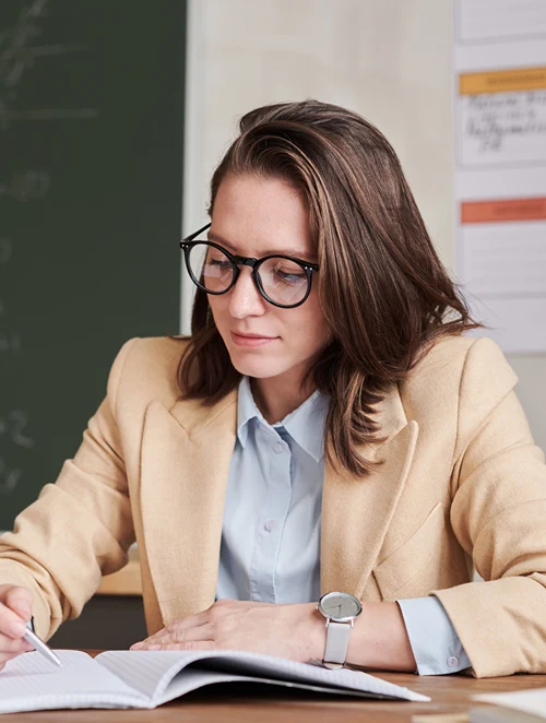 Professionally dressed female teacher writing at desk in her classroom.