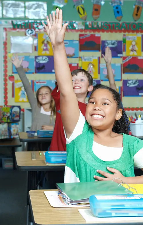 Middle school aged kids in row of desks eagerly holding up their hands in class.