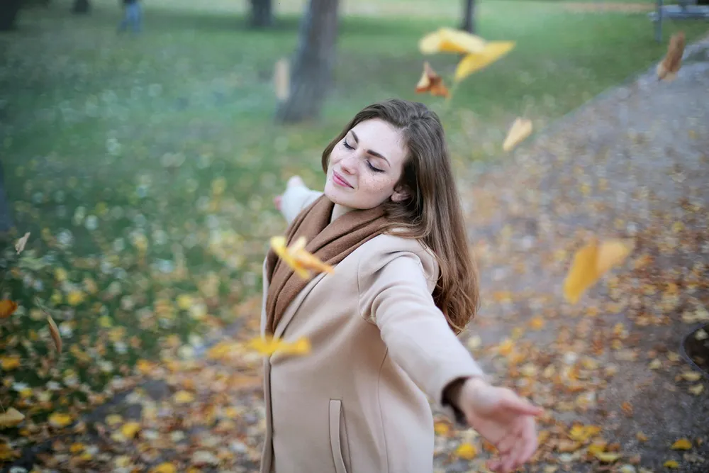 A woman in a park enjoying falling leaves with eyes closed and arms wide open.