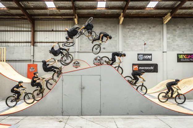 Sequence photo of a BMX rider performing a jump and mid-air trick inside an indoor skate park.