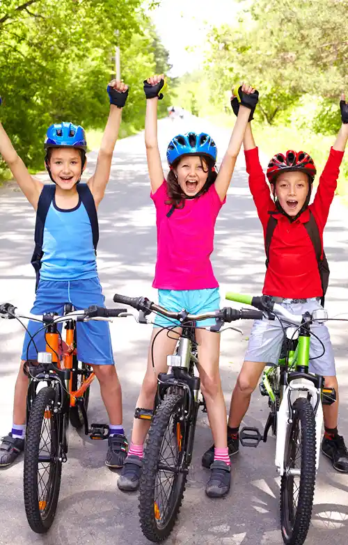 Three children wearing helmets riding bikes outdoors with their arms raised, smiling on a sunny day.