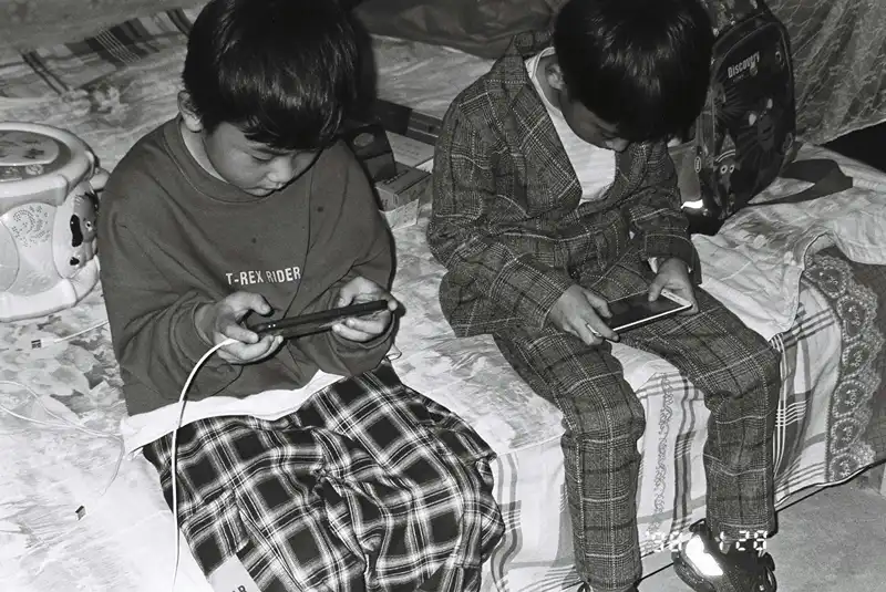 Two boys sitting on side of bed engaged in playing games on their phones.
