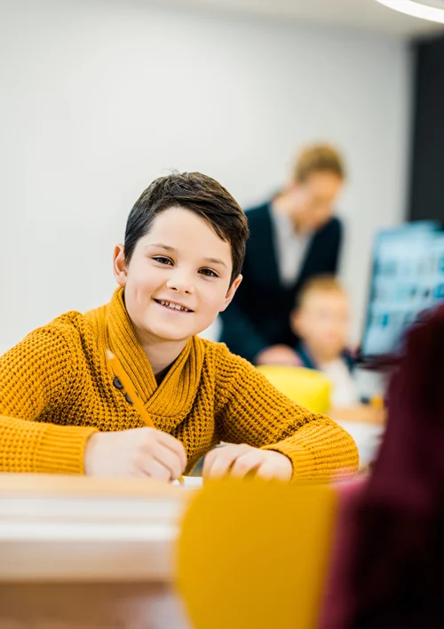 A young boy looks up from writing in his class.