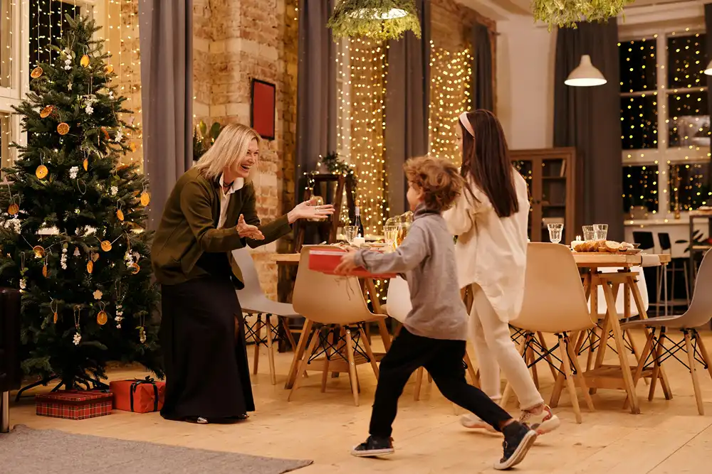 A mom welcomes her children with open arms in a larger room decorated for Christmas.