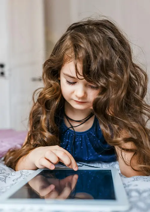 A little girl reads a book on a tablet on her bed