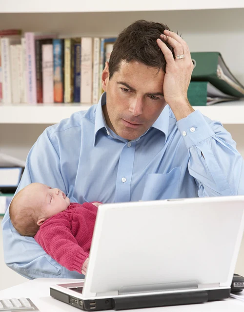 Stressed dad works on his laptop while holding a baby.
