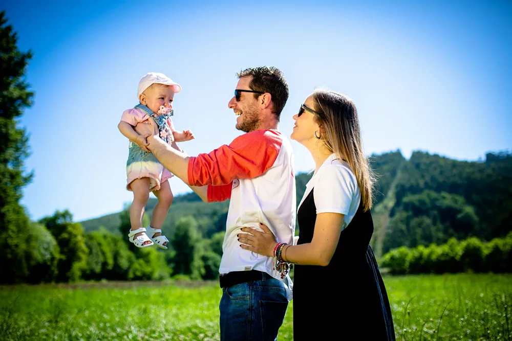 A father stands with mother as he holds up their baby in a green meadow.