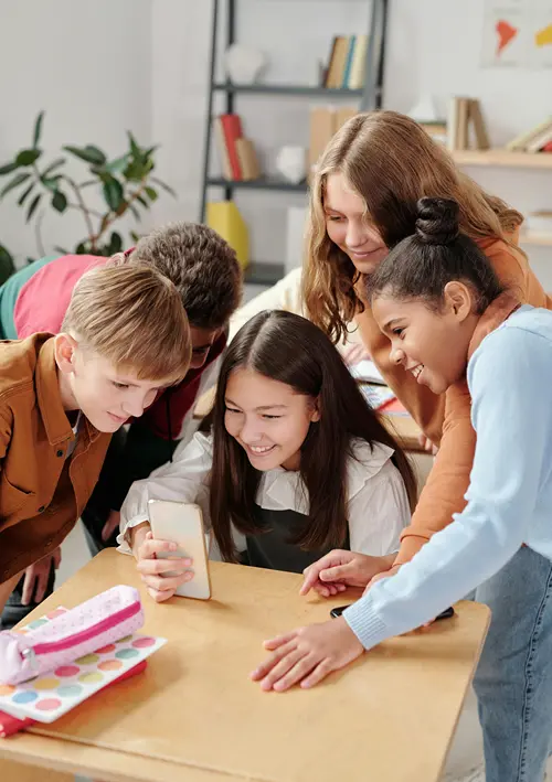 Children gathered around a girl seated at her classroom desk and smiling as they all look at her smartphone.