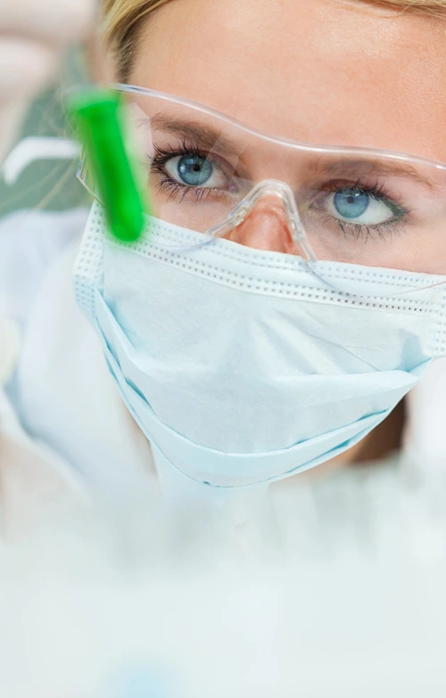 A female research scientist with mask looks closely at a test tube.